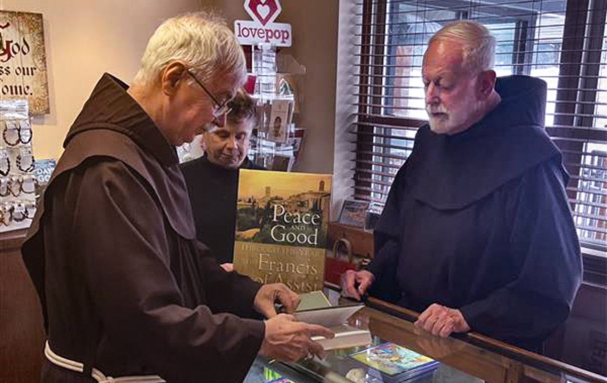 Two Franciscan friars and a woman stand at the counter of a Catholic gift shop. One of the friars is reading a book while the others look on.