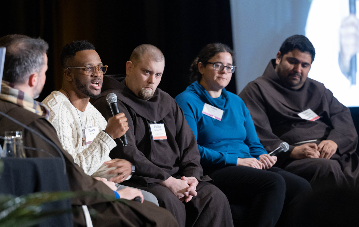 Five people sit on a stage. Two are holding microphones and three are wearing friar habits.