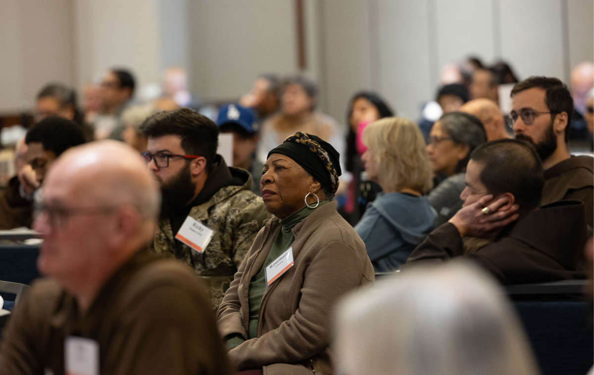 A woman sits among a crowd of people. She is wearing a black cap, hoop earrings and a brown jacket.