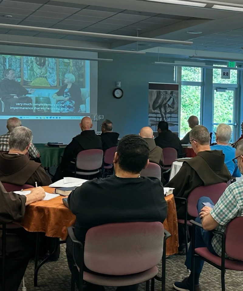 A group of men sit at tables watching a program on a screen. Some men are wearing friar habits.