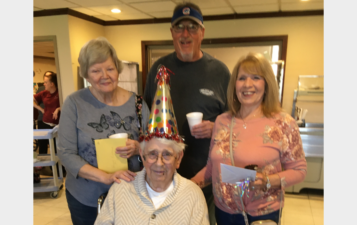 Two smiling woman and a man stand behind a friar wearing a festive birthday hat who is seated in a chair.