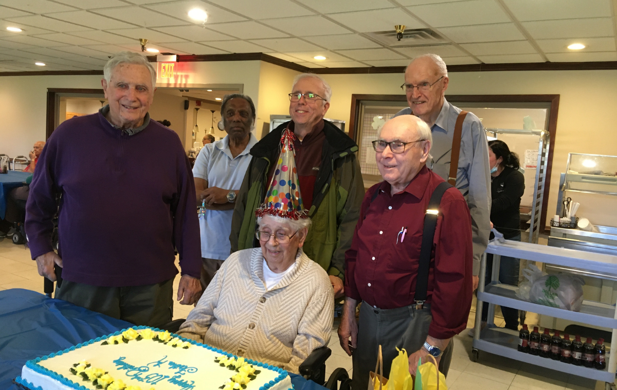 Five smiling men stand behind a happy man who is seated in front of a birthday cake. He is wearing a festive birthday hat.