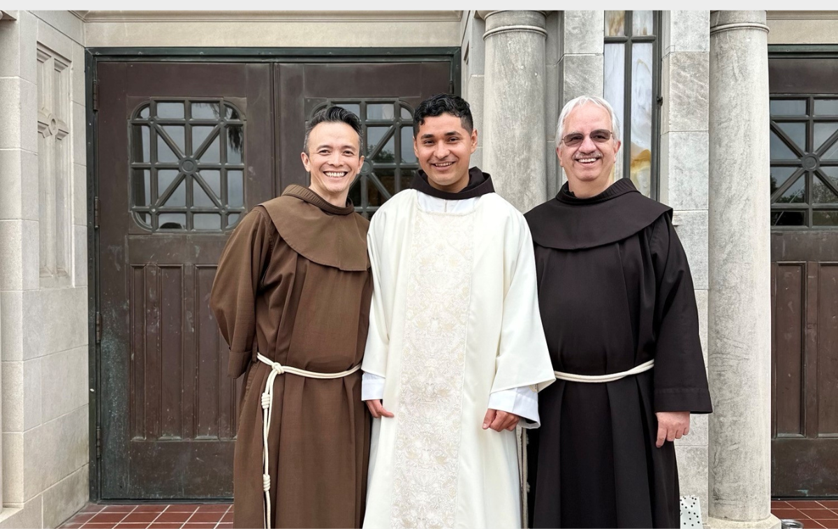 Two Franciscan friars wearing brown robes stand next to a friar who is wearing the white vestments of a deacon. All three men are smiling.