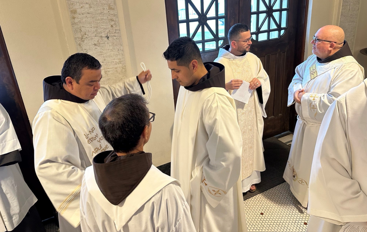 Franciscan friars wearing white vestments over their habits stand in the vestibule of a church.