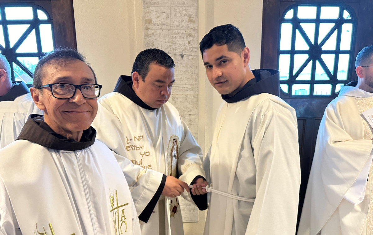 One friar helps another friar tie a cord around the white vestments he is wearing.