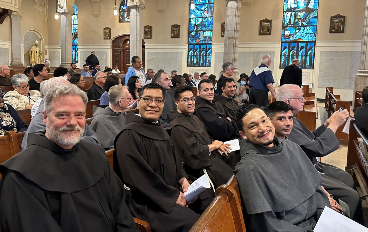 OFM and OFM Conventual friars smile for the camera as they sit in the pews of a church.