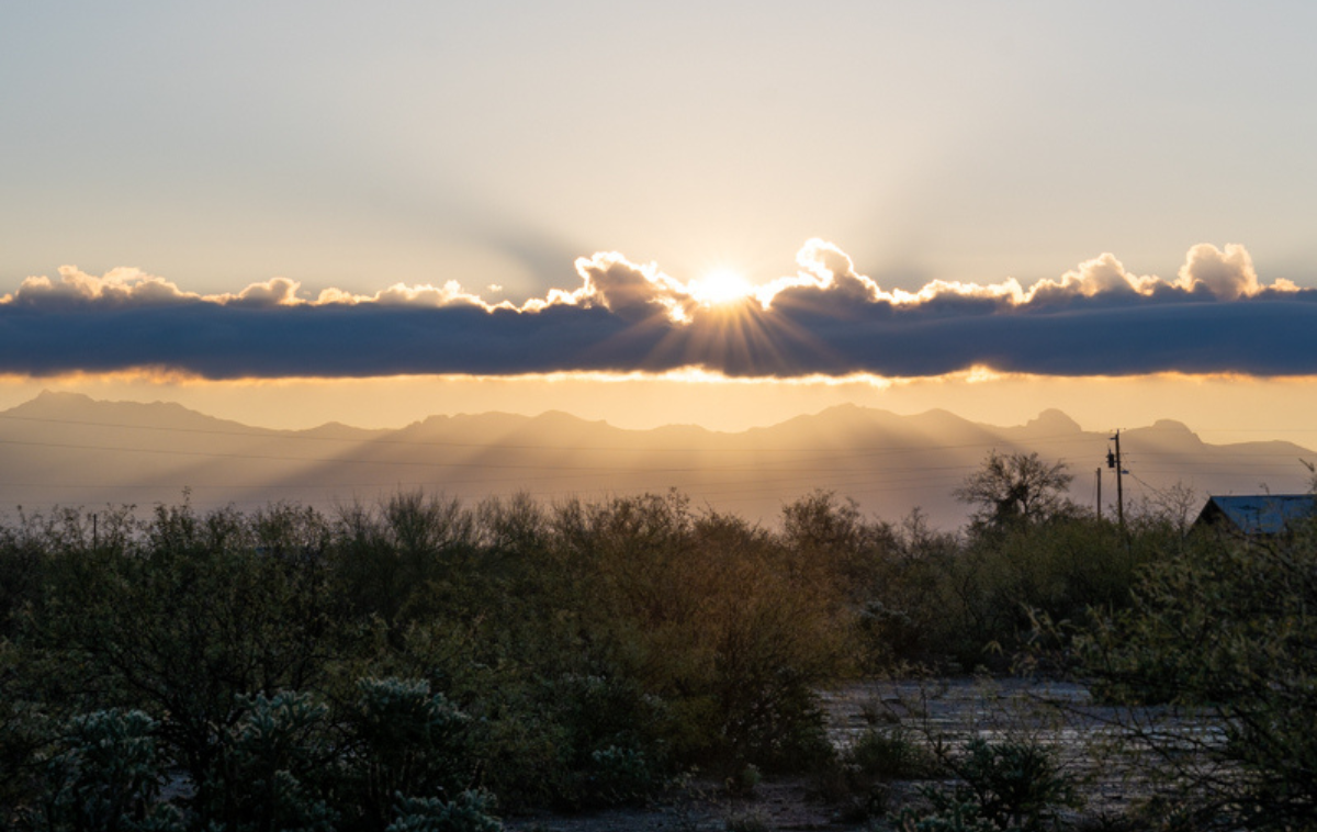Sun rays break through a line of dark purple clouds hovering over the desert. Mountains fade in and out of the haze on the horizon.