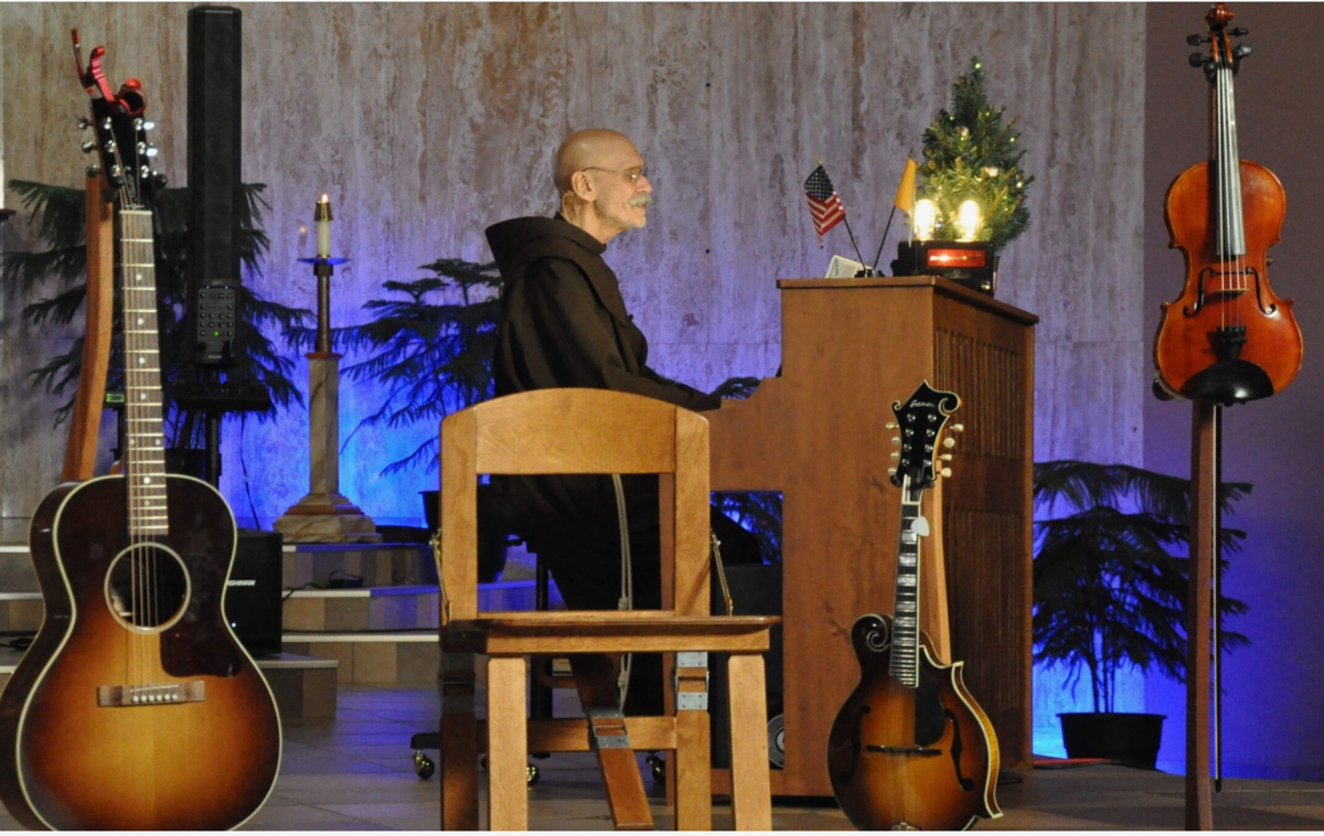 A friar plays the piano on a stage. Two guitars and a violin are placed on stands next to him.