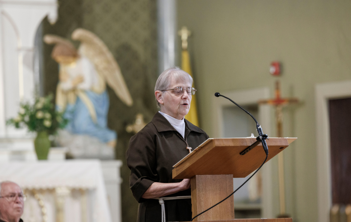 A woman wearing a brown Franciscan habit and a white rope cord with three knots wrapped around her waist reads from a lectern at the front of a church.