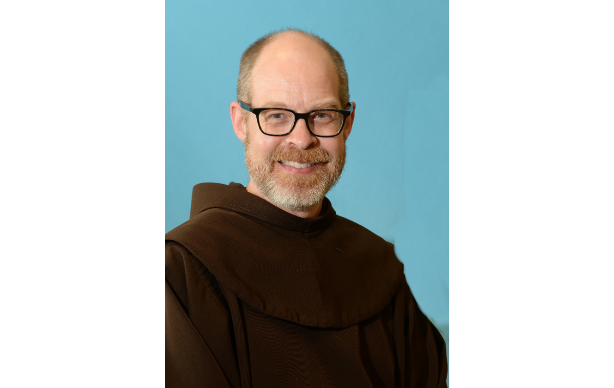 A bearded man wearing glasses and smiling, wearing a friar habit.