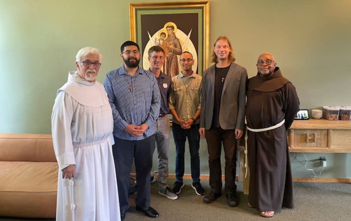 Six men pose for a picture i a room between a couch and a table and in front of an image of a saint on the wall.