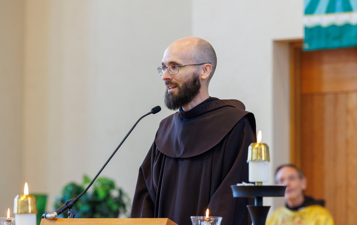 A friar in a brown habit kneels before a priest wearing yellow vestments. The priest holds the hands of the friar as he reads from a book.