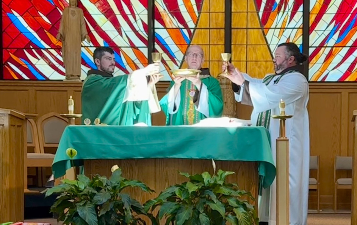 Four men stand in a room beside a plant and in front of a crucifix. There is a statue of Jesus Christ behind them.