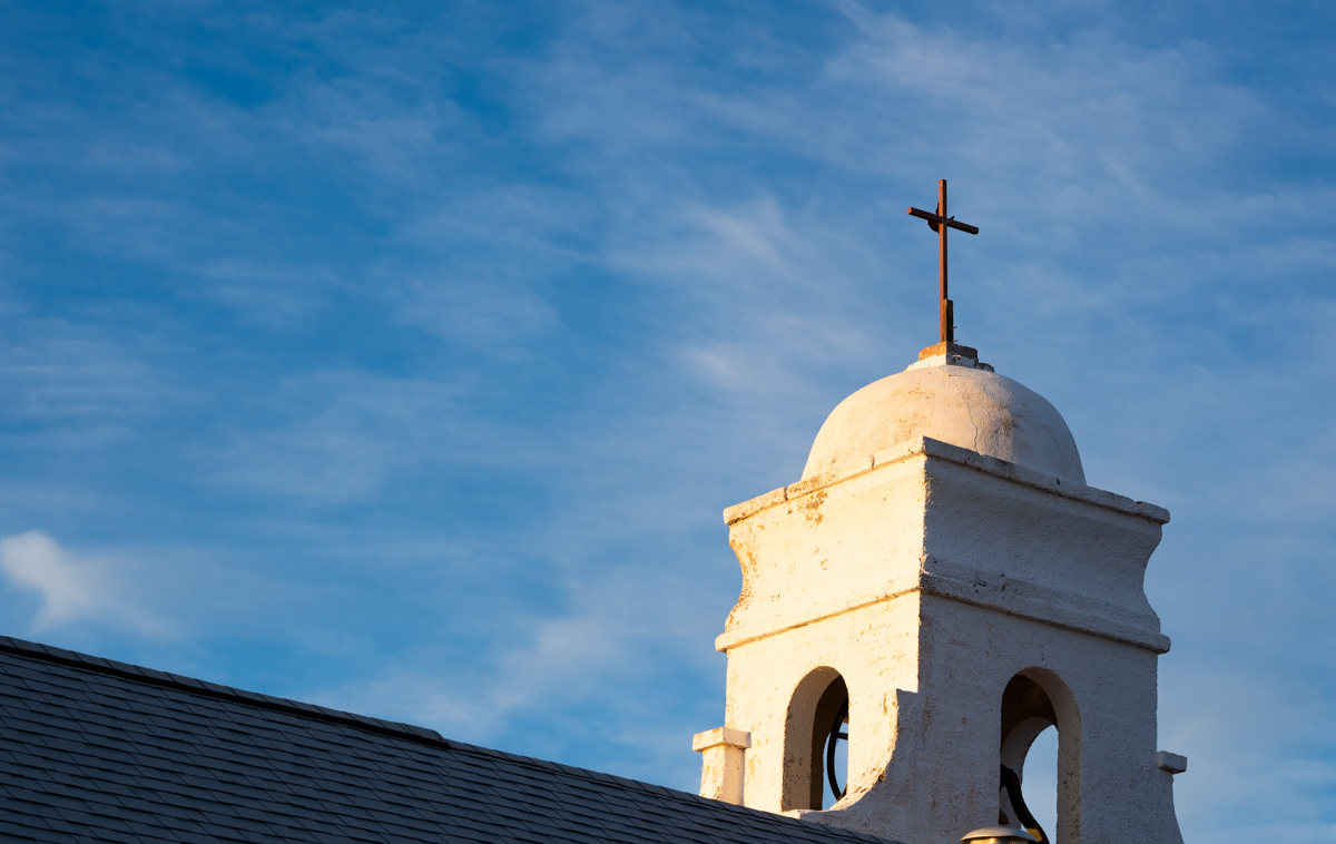 The steeple of a small white mission-style church glows in the sun against a blue sky