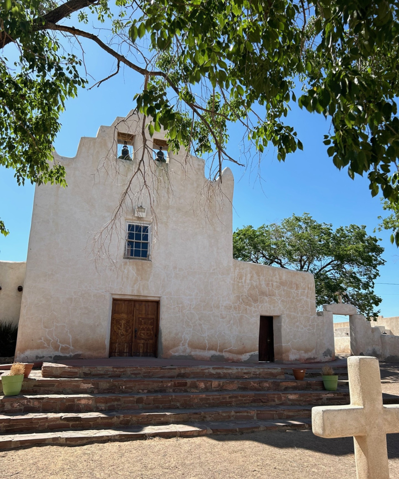 An old mission-style church with two small bells in its steeple stands proudly against the bright blue sky.