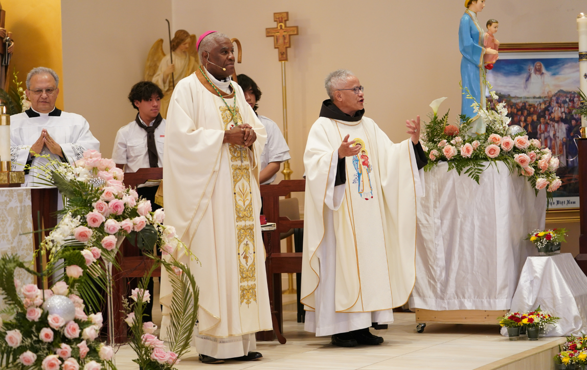 Standing in front of an altar, Br. David Phan greets parishioners. He stands to the left of a statute of Our Lady of La Vang that is decorated with flowers. Next to him is Bishop Jacques Fabre-Jeune, CS.