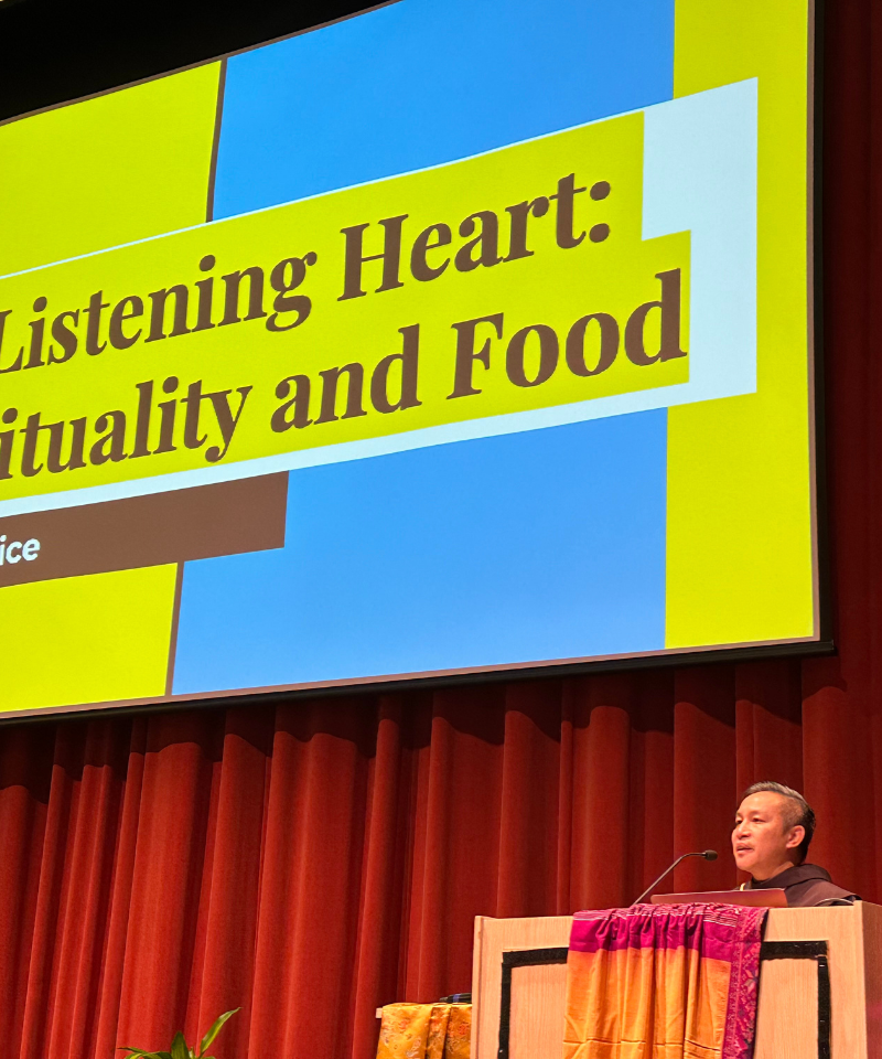 A man stands behind a wooden podium next to a screen that says A listening heart spirituality and food.