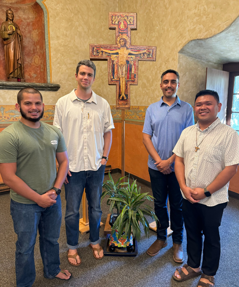 Four men stand in a room beside a plant and in front of a crucifix. A statue of Jesus Christ is behind them.
