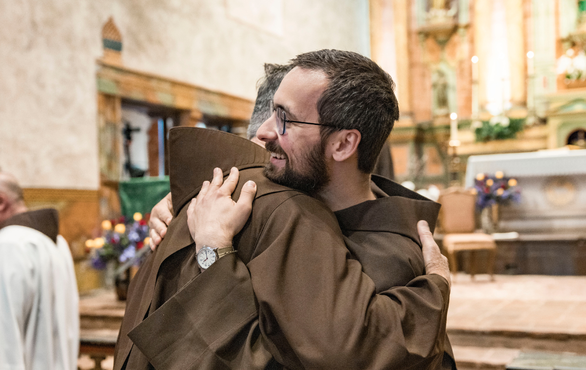 A smiling younger friar receives the sign of peace from an older friar.