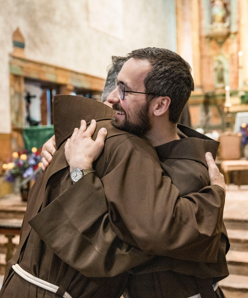 Two friars smile as they share a hug before an altar of a Spanish-style mission church.