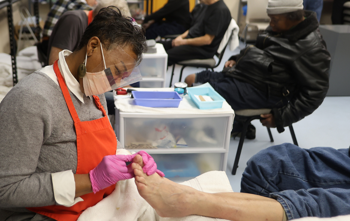 A smiling woman wearing a red apron and pink gloves over her clothes trims the nails of an outstretched foot.