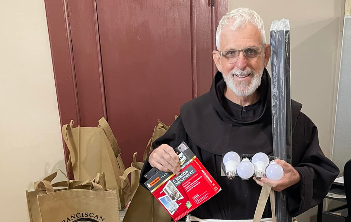 A smiling friar holds a window insulation kit and four lightbulbs.