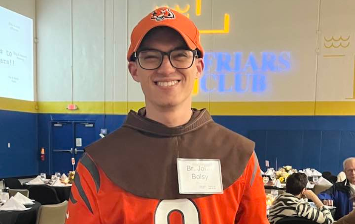 A young friar smiles at the camera. He is wearing a Cincinnati Bengals jersey and hat over his Franciscan habit.