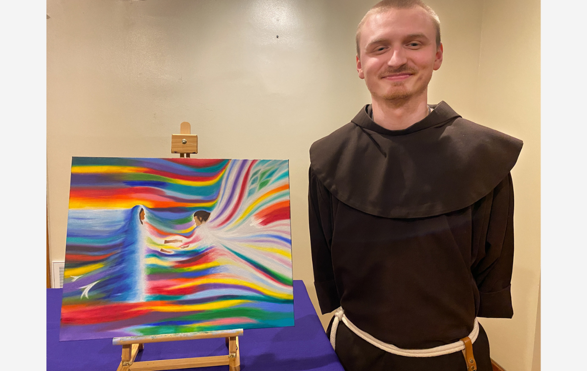A friar in his 20s smiles as he stands next to a medium-sized rectangle painting. The painting is of Mary receiving the news that she will be the mother of God's son from the angel Gabriel.