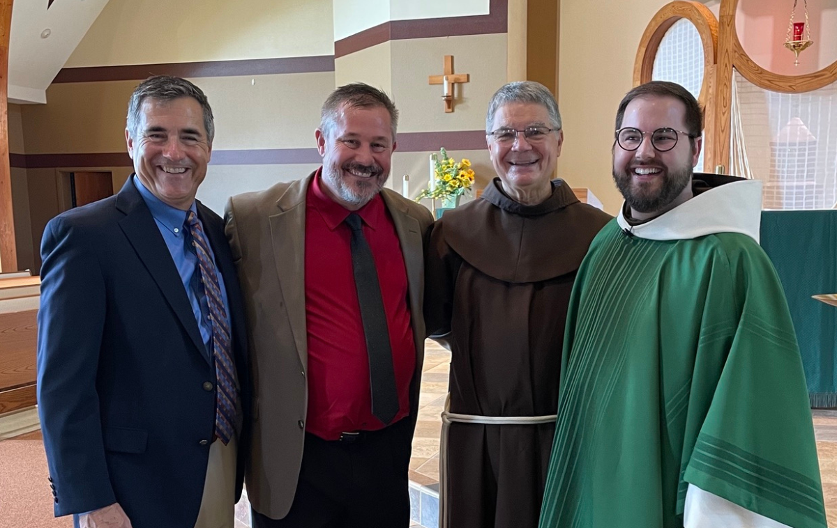 Two men wearing suits and ties stand next to a Franciscan friar in a brown habit and a priest in green vestments. All are smiling. You can feel their joy.