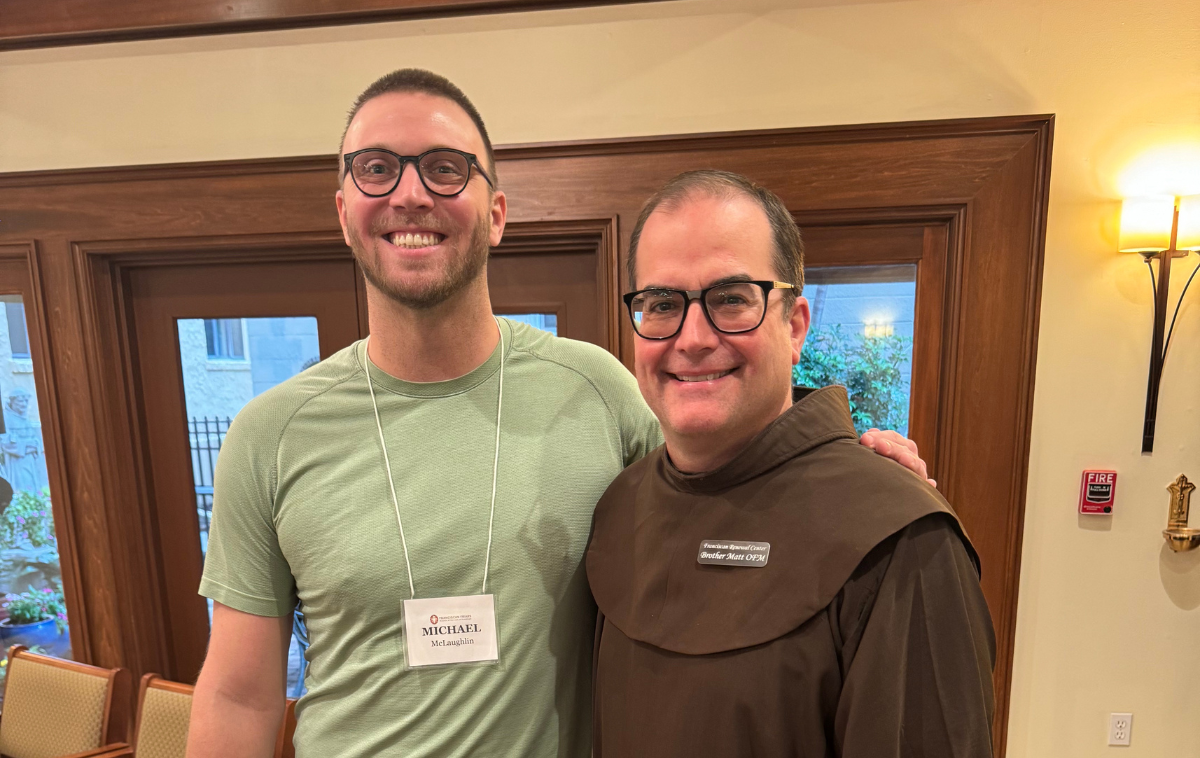 A friar stands next to a man in street clothes. Both are smiling.