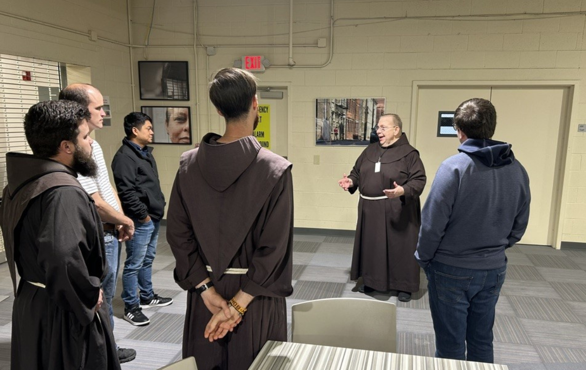 A friar speaks to a group of six men in the vestibule of a soup kitchen.