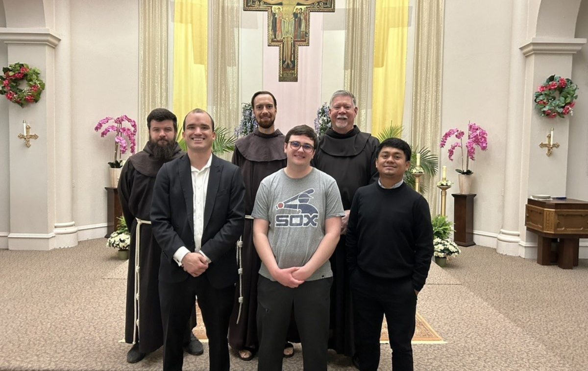 Three friars dressed in their brown habits and three men stand smiling in front of an altar.