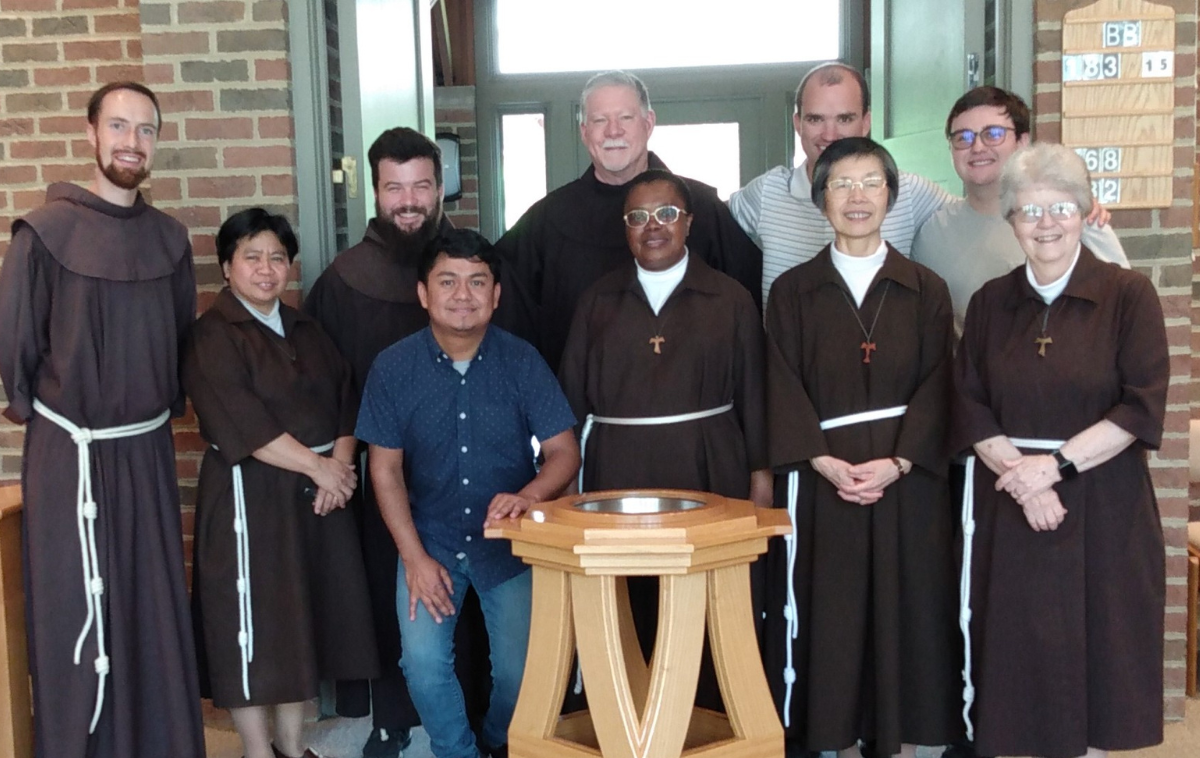 Three friars in habits, three men in street clothes, and four Poor Clares stand smiling behind a baptismal font