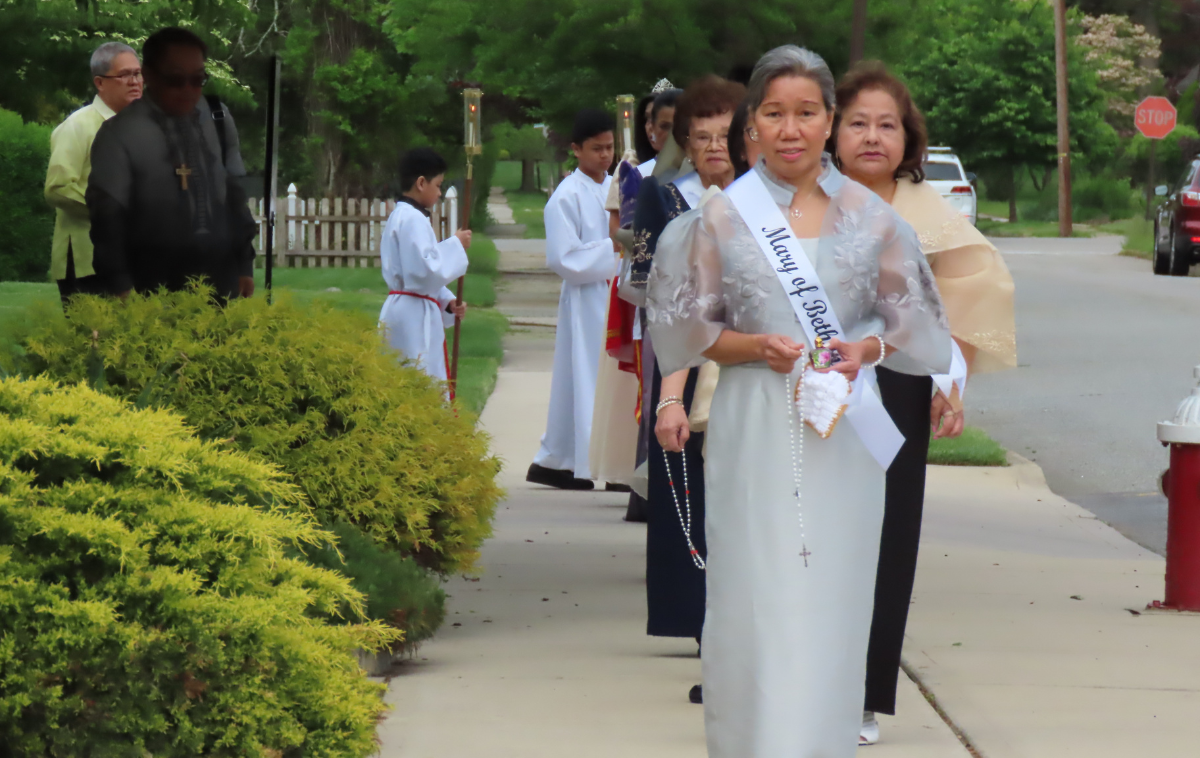 A group of women in formal wear process down a residential street. They carry rosaries and are wearing sashes representing different women from the Bible and Church history.