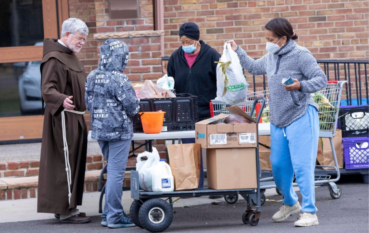 A friar welcomes three people who are picking up bags of food.