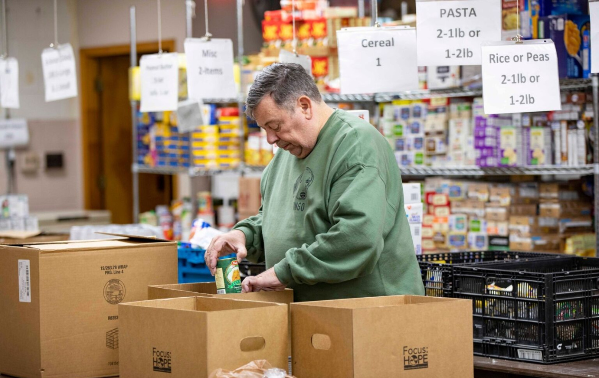 A man wearing a green sweatshirt unpacks boxes of canned goods.