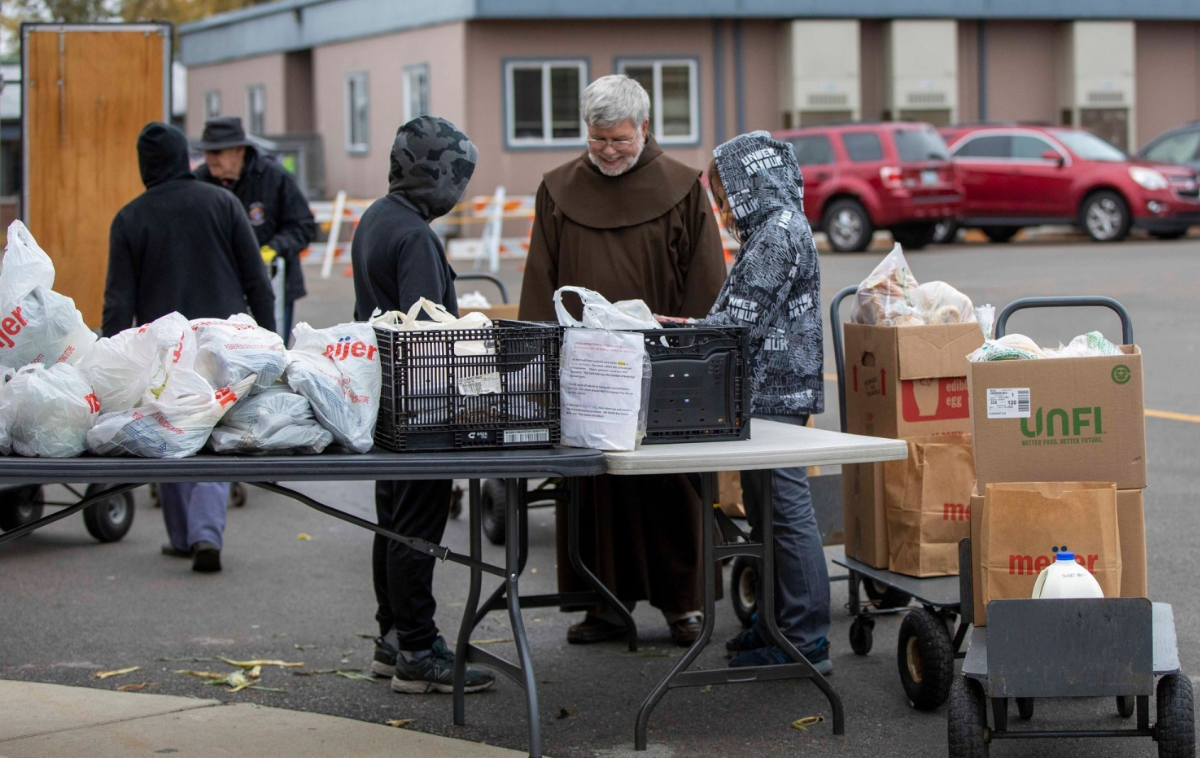 A friar greets people who are picking up bags of food at a long folding table.