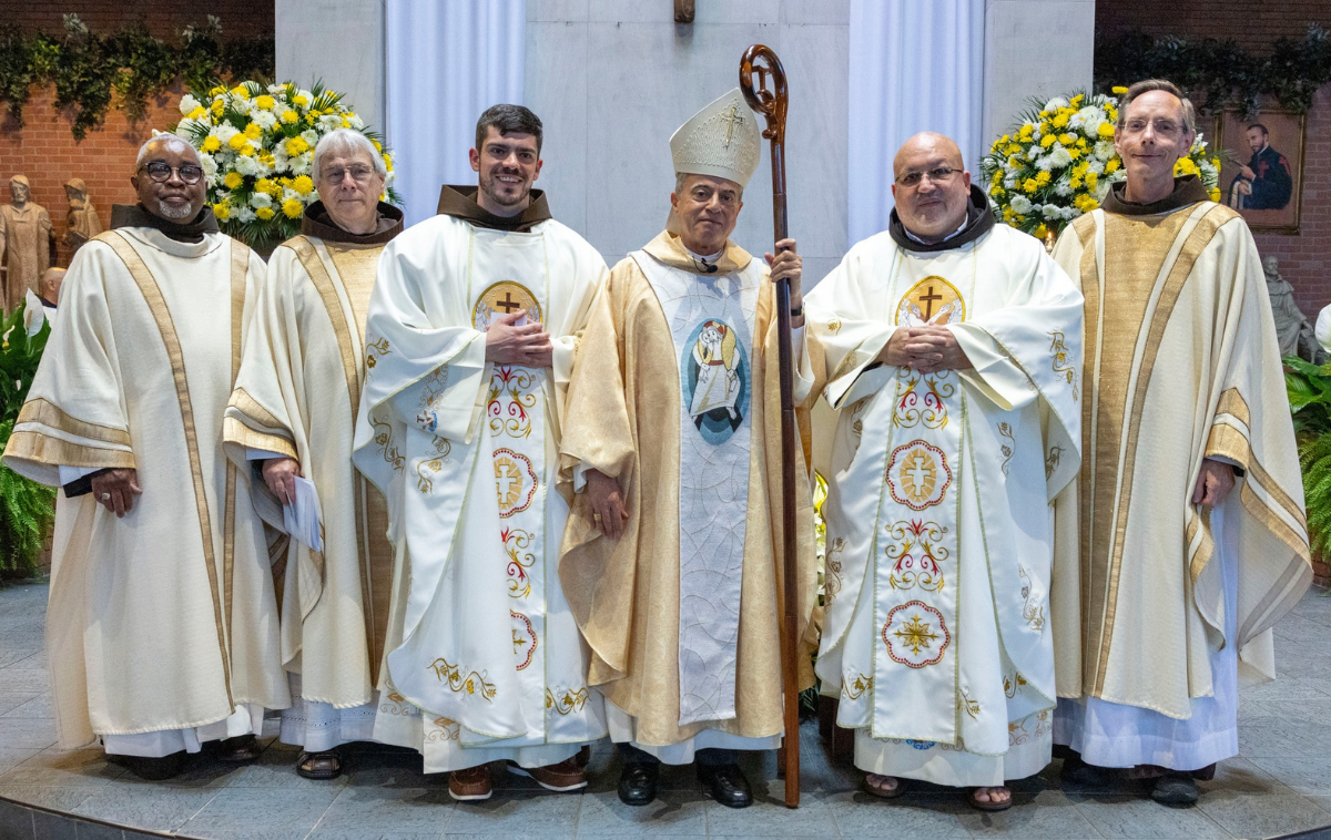A permanent deacon, four priests and an archbishop stand before an altar and smile at the camera