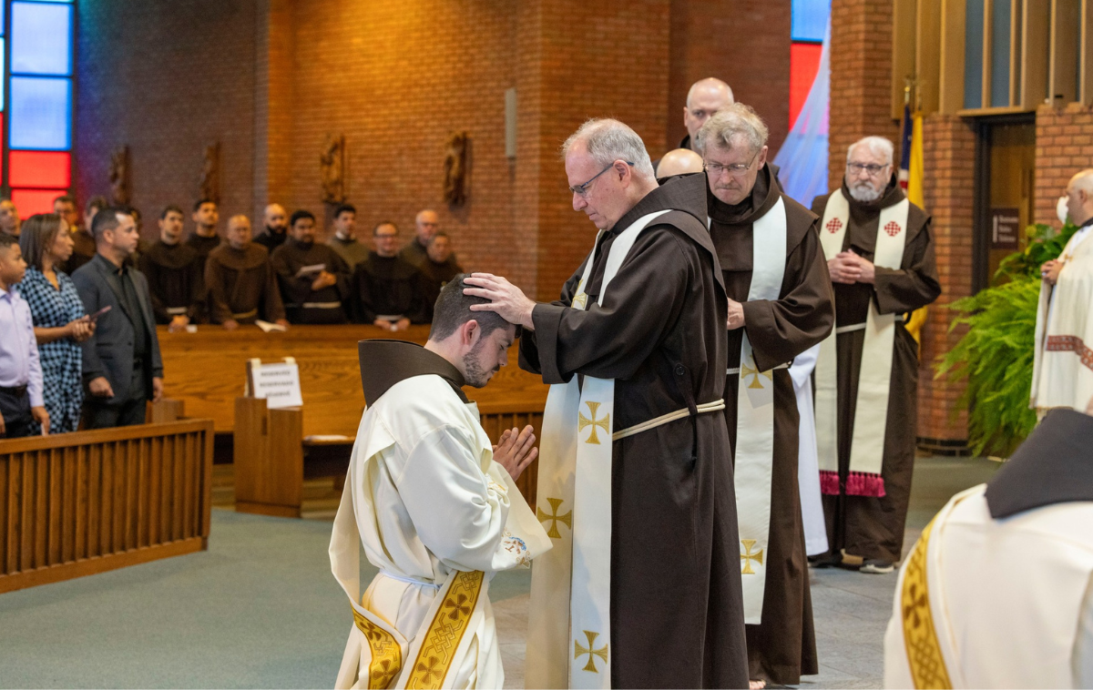 A newly ordained priest kneels for a blessing from a friar