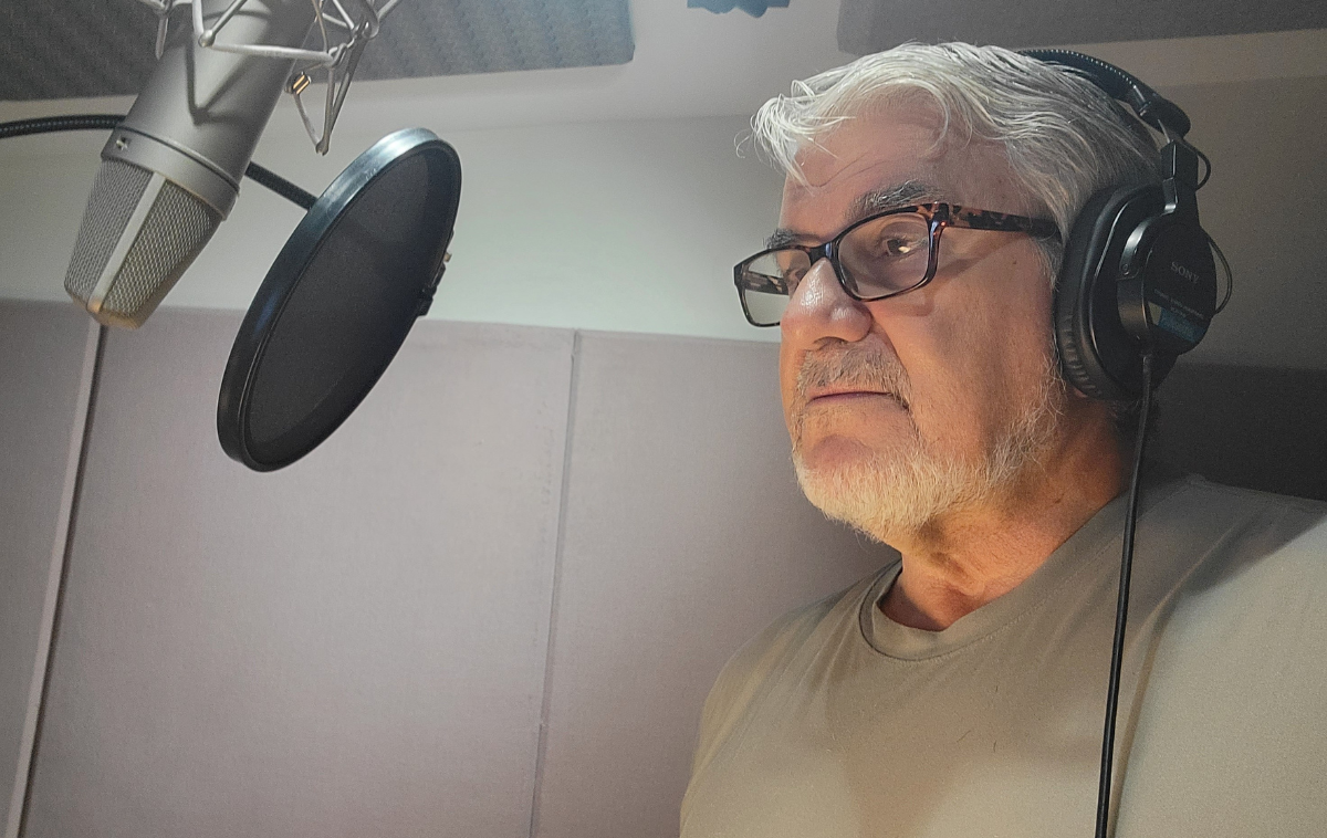 A friar with a close-cropped beard wearing headphones stands in front of a microphone in a recording studio