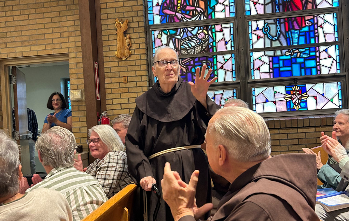 A friar standing in the pews of a crowded church lifts his hand to acknowledge the people around him, who are clapping for him.