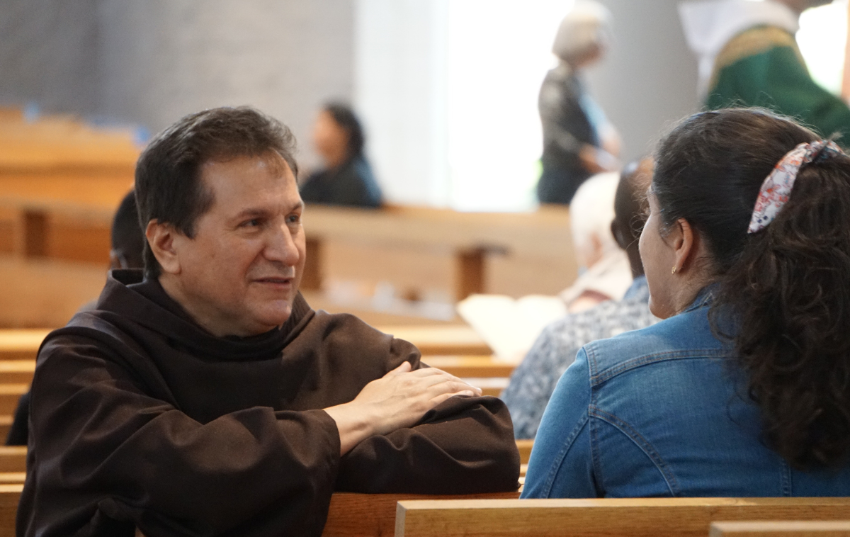 A friar sits and listens to a woman in the pews of a church.