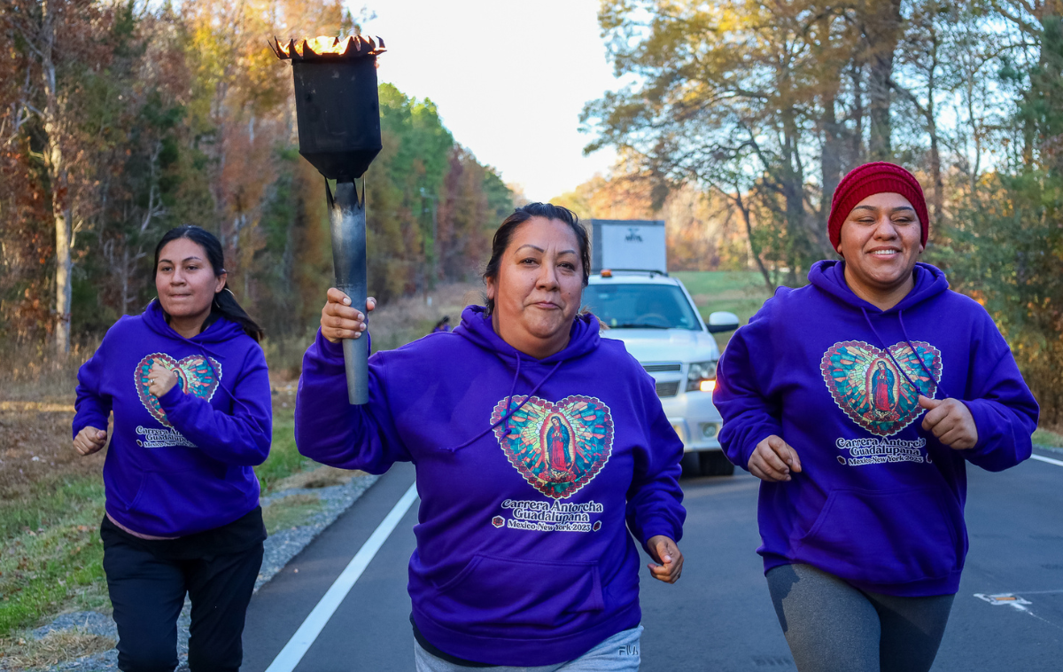 Three people jog down a road. One is holding a burning torch. All three are wearing shirts that say 