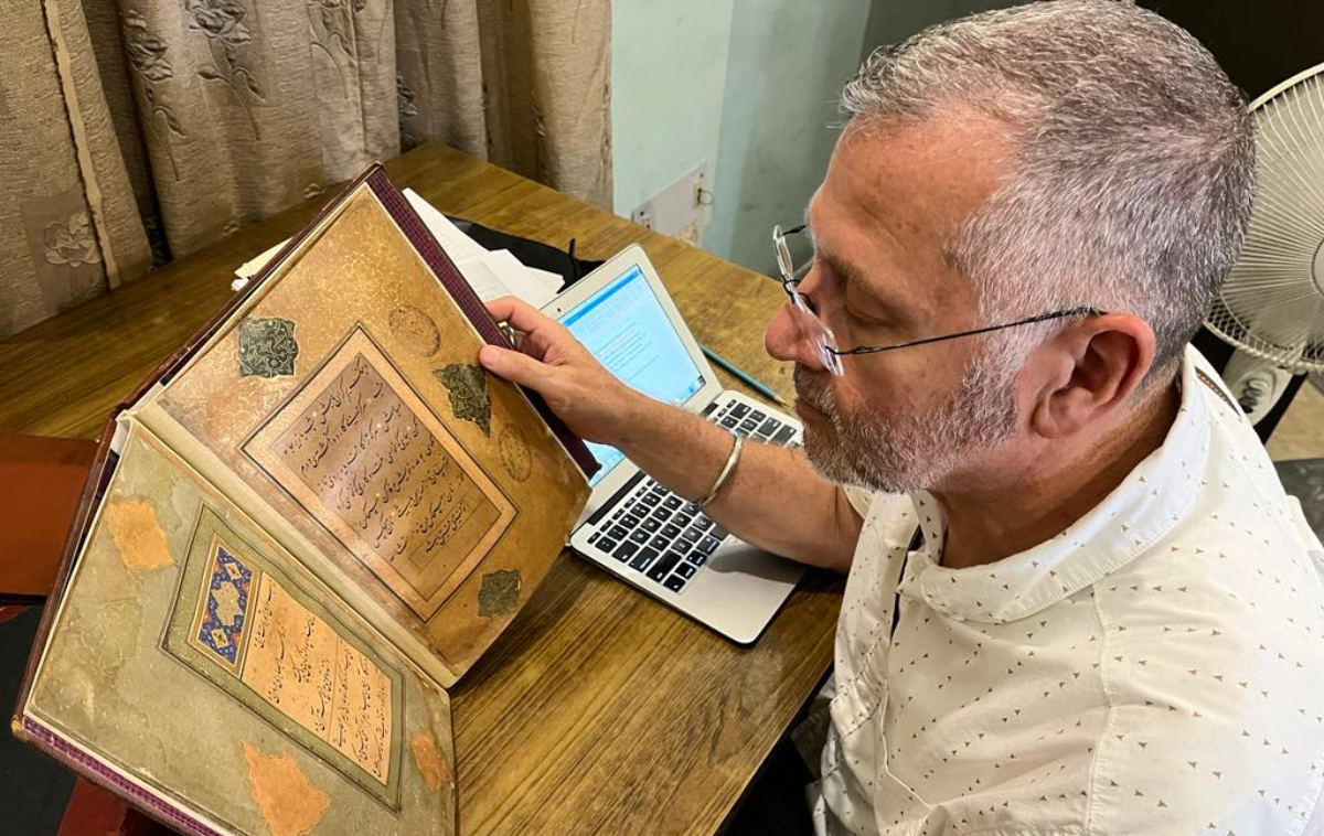 A man studies a medieval manuscript. There is Arabic script on the page, bordered by intricate artwork.