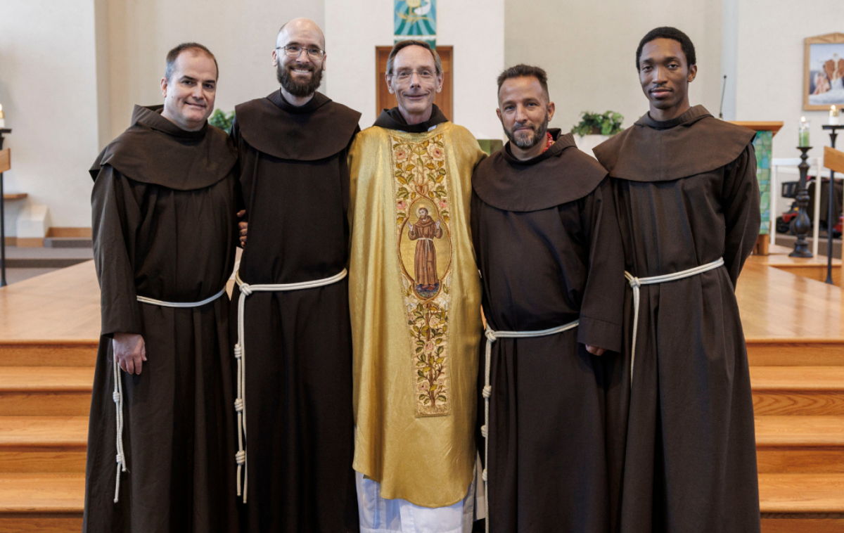 Four men stand in a room beside a plant and in front of a crucifix. There is a statue of Jesus Christ behind them.