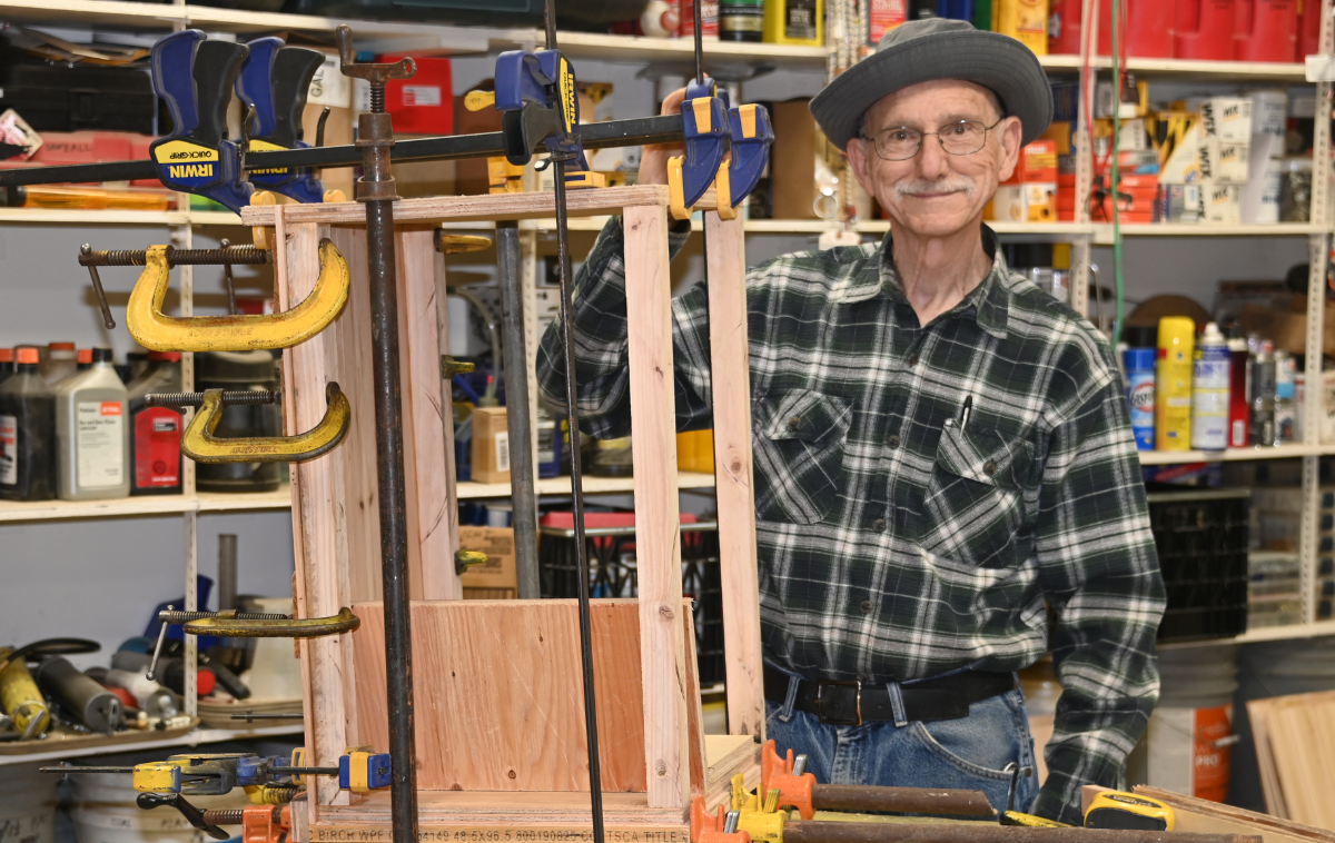 A man wearing a green and white plaid shirt stands next to a cabinet he is building.