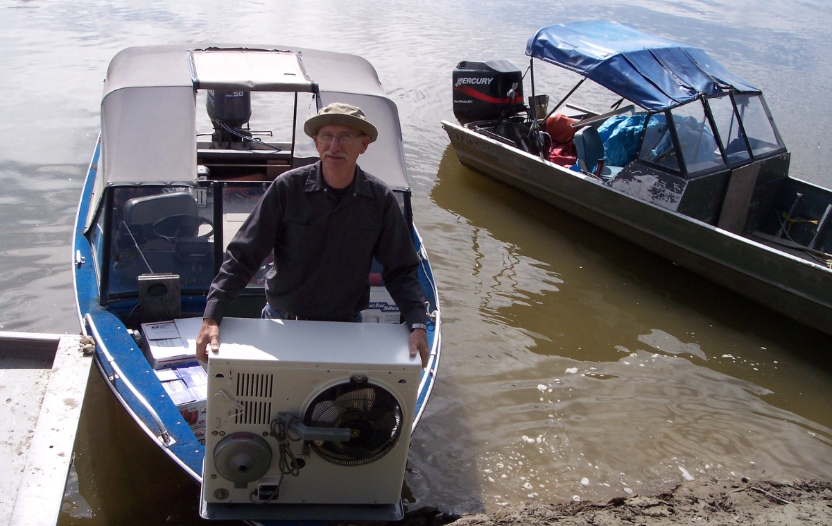 A man wearing a fisherman's hat stands at the helm of a small boat that is docked on the Yukon River..