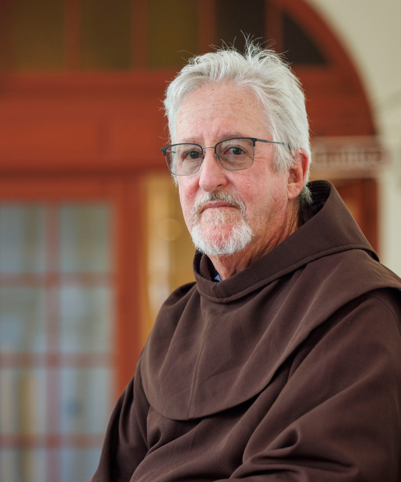 A bearded man wearing glasses sits outside wearing a friar habit.