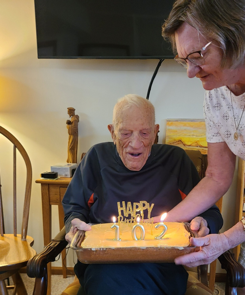 A man sitting in a chair is given a birthday cake by a woman standing next to him.