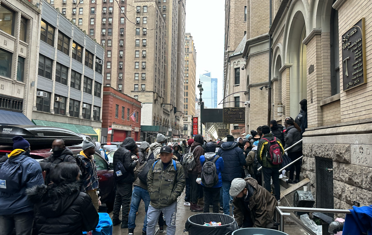 Dozens of people in winter clothing line up along a sidewalk and onto a staircase as they wait outside a building on a city street.